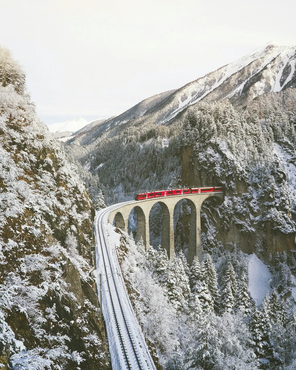 Train crossing a curved stone viaduct through snowy mountains